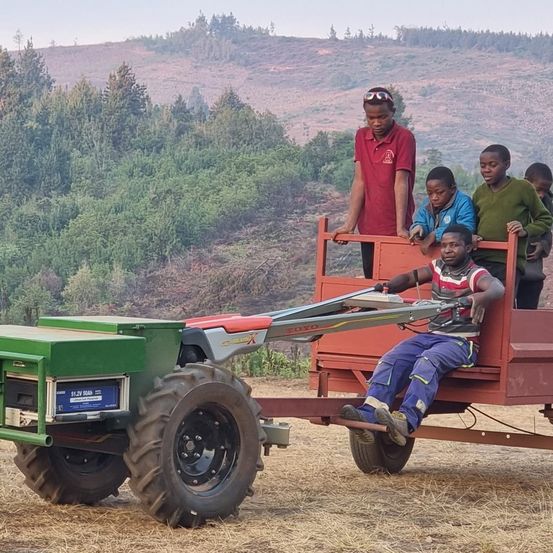 Ein Mann fährt einen roten Traktor mit Anhänger, auf dem mehrere Kinder sitzen und am Rand stehen. Dahinter steht ein Junge mit einer Schutzbrille. Sie befinden sich in einem hügeligen Gebiet mit grünen Bäumen.