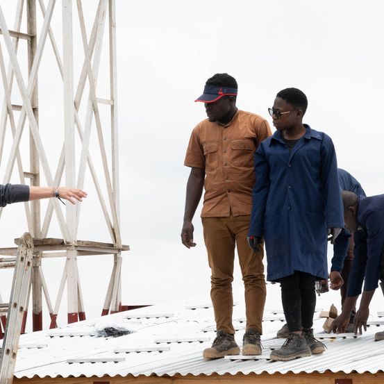 Eine Gruppe von Menschen, darunter ein Mann in einem braunen Hemd und einer in einem blauen Mantel, steht auf einem Metalldach. Ein Mann im Hintergrund beugt sich nach unten. Eine andere Person streckt ihren Arm von links aus. Ein Metallgerüst steht hinter ihnen.