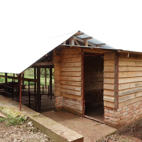 Ein kleines Holzhaus mit einem Metalldach und einer offenen Tür. Der Bau ist teilweise mit Ziegeln errichtet. Die Umgebung zeigt einen offenen Bereich mit etwas Vegetation.