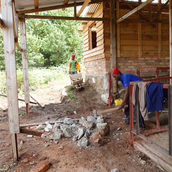 Zwei Männer arbeiten auf einer Baustelle. Einer schlägt mit einem Hammer auf Steine ein, während der andere einen Schubkarren schiebt. Ein Holzhaus im Hintergrund.