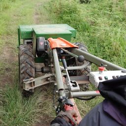 Ein grüner Traktor befindet sich auf einem Grasweg, wobei eine Person ein Werkzeug in der Hand hält. Der Traktor hat einen grünen Körper, schwarze Reifen und eine orangefarbene Vorderseite.