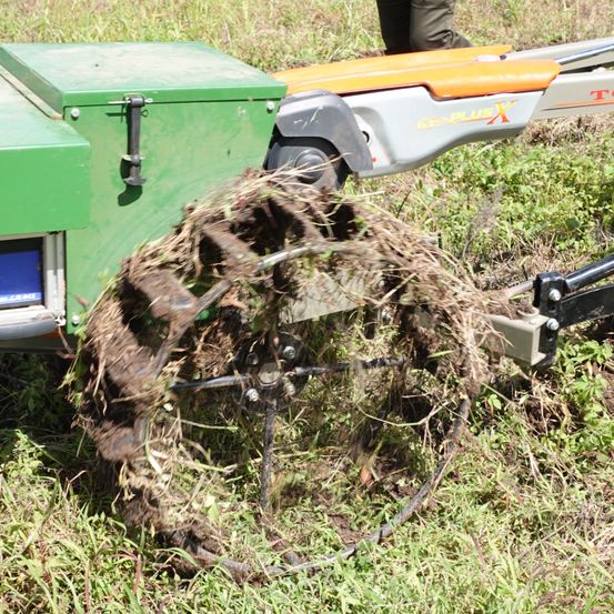 Eine grüne Maschine mit einem Rad verarbeitet Gras auf einem Grasfeld. Eine Person steht in der Nähe und beobachtet.