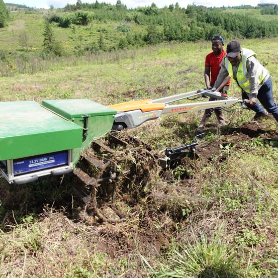 Zwei Personen bedienen eine grün-orange Maschine mit dem Wort 'TOYO' auf einem Grasfeld. Eine Person trägt eine reflektierende Weste.