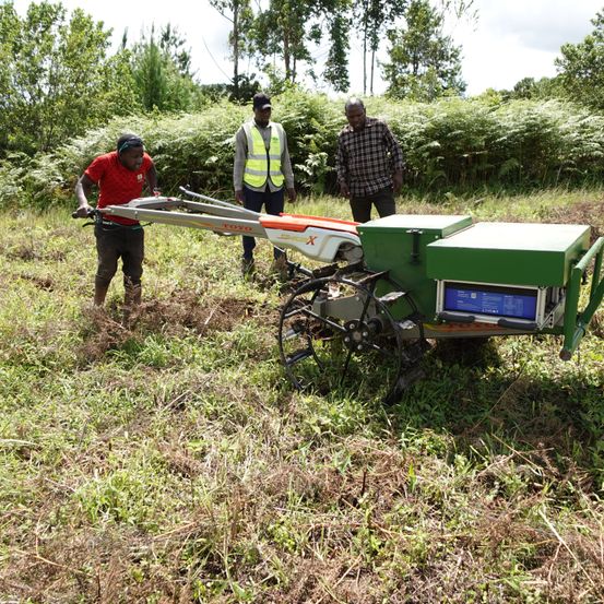 Drei Männer bedienen einen grünen Traktor auf einem Grasfeld mit Bäumen im Hintergrund.
