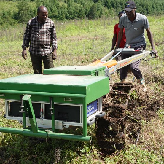Zwei Männer benutzen einen grünen Maschinenwagen, um Erde in einem Feld zu graben. Das Fahrzeug hat einen Markennamen, 'TOYO'. Die Maschine gräbt die Erde in Reihen. Ein anderer Mann steht in der Entfernung.