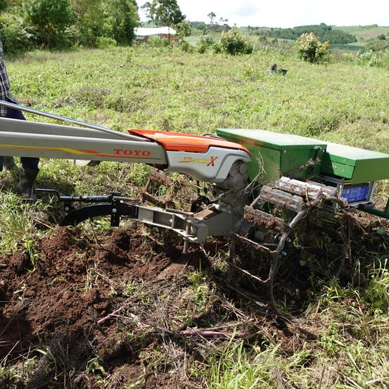 Eine Person verwendet einen roten und grünen Traktor, mit der Aufschrift 'TOYO', um Erde auf einem grün bewachsenen Feld zu pflügen.