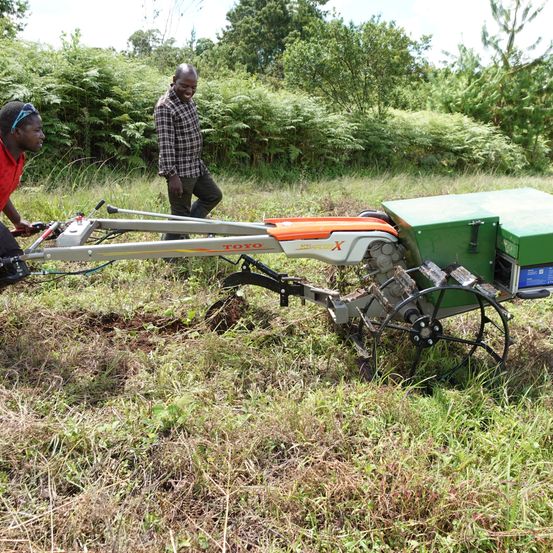 Zwei Männer benutzen eine grün-orange Maschine in einem Grasfeld, einer lächelt und der andere schaut auf die Maschine.