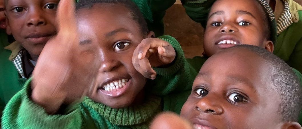 Eine Gruppe junger Kinder in grünen Uniformen lächelt und posiert für ein Foto mit erhobenen Fingern. Sie scheinen sich in einem Klassenzimmer zu befinden.