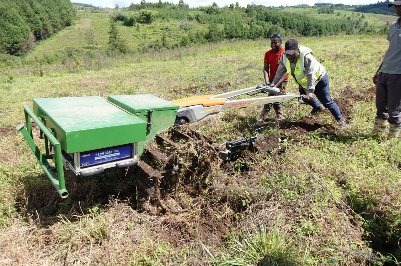 Zwei Männer benutzen einen großen Traktor, um den Boden auf einem Feld zu pflügen. Ein Mann trägt eine reflektierende Weste und der andere trägt ein rotes Hemd.