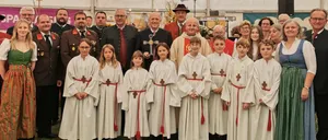 Eine Gruppe von Menschen, darunter Kinder in weißen Gewändern und Erwachsene in traditioneller Kleidung, posiert für ein Foto unter einem Zelt mit dem Schriftzug 'Leitgeb Festzeltverleih'.