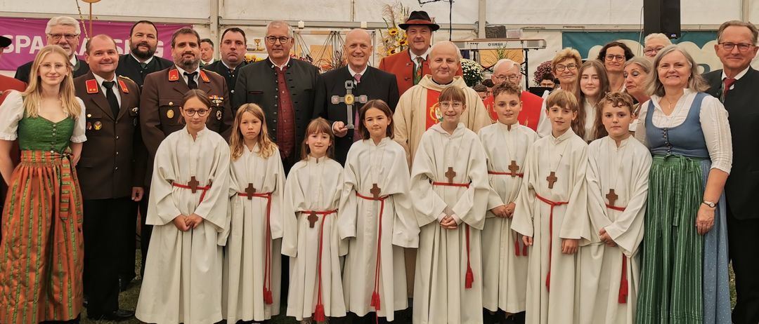 Eine Gruppe von Menschen, darunter Kinder in weißen Gewändern und Erwachsene in traditioneller Kleidung, posiert für ein Foto unter einem Zelt mit dem Schriftzug 'Leitgeb Festzeltverleih'.