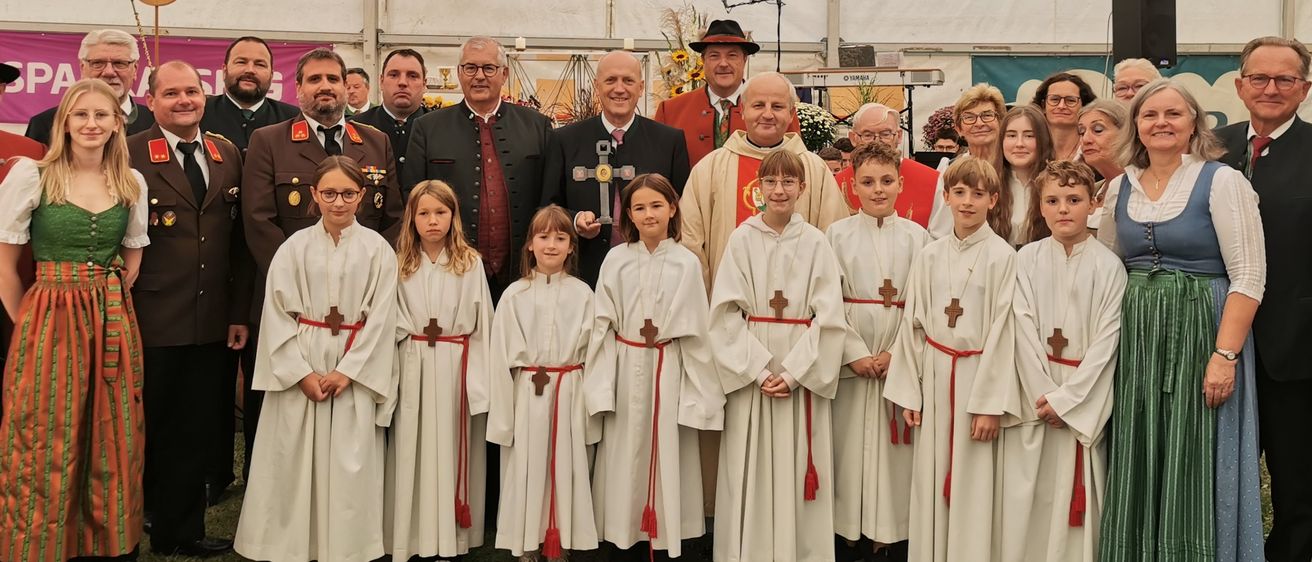 Eine Gruppe von Menschen, darunter Kinder in weißen Gewändern und Erwachsene in traditioneller Kleidung, posiert für ein Foto unter einem Zelt mit dem Schriftzug 'Leitgeb Festzeltverleih'.