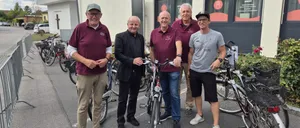 Fünf Männer stehen vor einem Gebäude mit Fahrrädern, zwei halten ihre Räder. Sie sind lässig gekleidet, einer trägt einen Hut und ein anderer einen schwarzen Anzug.