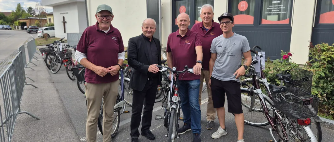 Fünf Männer stehen vor einem Gebäude mit Fahrrädern, zwei halten ihre Räder. Sie sind lässig gekleidet, einer trägt einen Hut und ein anderer einen schwarzen Anzug.