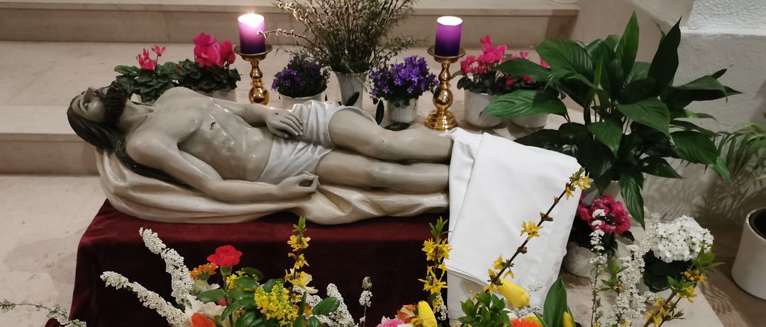 Eine Statue von Jesus liegt auf einem Tisch, der mit einem weißen Tuch bedeckt ist. Um die Statue herum befinden sich verschiedene Blumen und Pflanzen, darunter eine Vase mit Blumen und eine Pflanze mit rosa Blüten.