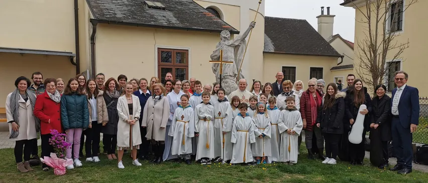 Eine große Gruppe von Menschen, einschließlich Kinder in weißen Gewändern, stehen vor einer Kirche mit einer Statue dahinter. Die Kirche hat ein grünes Dach und mehrere Fenster.