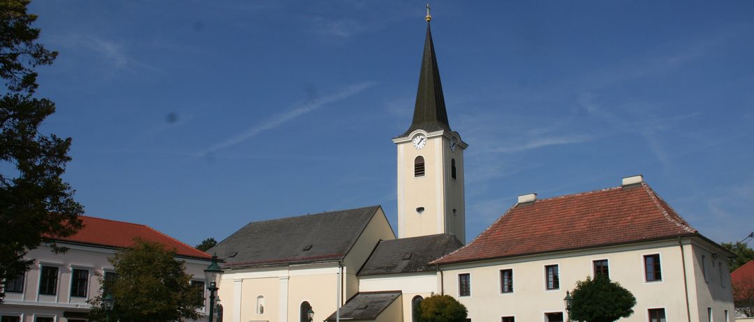 Eine beigefarbene Kirche mit einem Turm und einer Uhr steht vor einem gebäude mit rotem Dach. Eine große Felsstruktur steht vor der Kirche. Bäume und Büsche befinden sich um die Kirche herum.