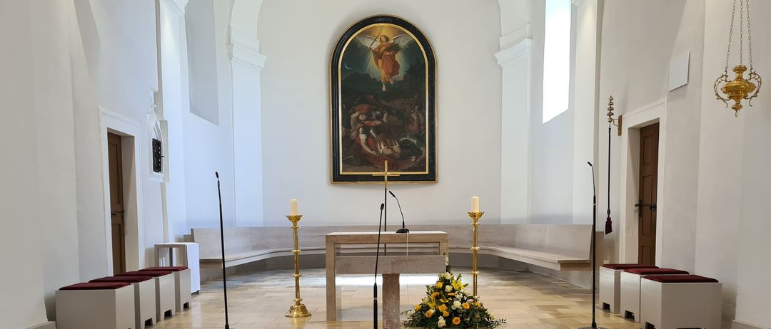 Ein Altar mit Kerzen und Blumen, in einer geräumigen, hellen Kirche. Ein gerahmtes Gemälde schmückt die Wand darüber, und Bänke flankieren den Bereich.