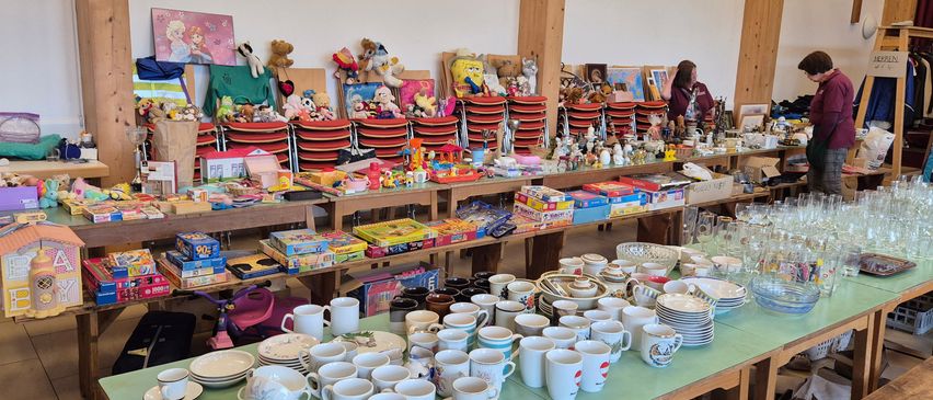 Ein großer Raum mit Holztischen und Regalen ist mit verschiedenen Gegenständen wie Tellern, Tassen, Spielzeug, Büchern und Kisten gefüllt. Zwei Frauen stehen im Hintergrund.