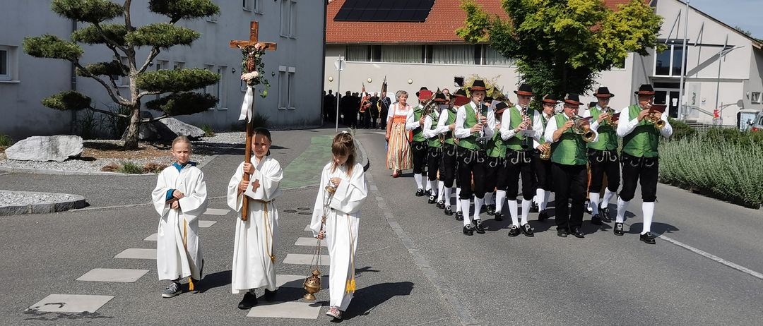 Eine Gruppe von Kindern und eine Blaskapelle sind in einem Umzug auf einer Straße. Die Kinder halten ein Kreuz und eine Kerze. Die Band marschiert in Reihe.