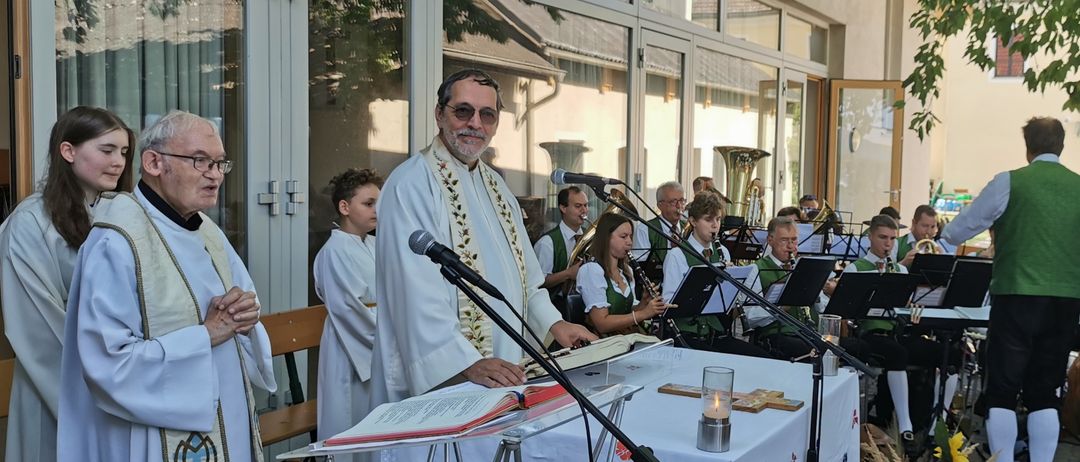 Ein Priester steht an einem Pult mit einem Mikrofon und liest aus einem Buch, während eine Band hinter ihm spielt. Die Umgebung ist im Freien, mit einem Gebäude und Bäumen im Hintergrund.