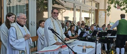 Ein Priester steht an einem Pult mit einem Mikrofon und liest aus einem Buch, während eine Band hinter ihm spielt. Die Umgebung ist im Freien, mit einem Gebäude und Bäumen im Hintergrund.