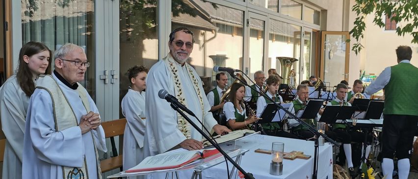 Ein Priester steht an einem Pult mit einem Mikrofon und liest aus einem Buch, während eine Band hinter ihm spielt. Die Umgebung ist im Freien, mit einem Gebäude und Bäumen im Hintergrund.