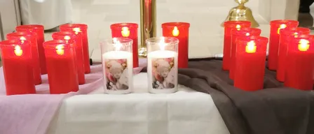 Ein Tisch mit weißer Tischdecke ist mit einem rosa und einem braunen Tuch bedeckt. Auf dem Tisch befinden sich mehrere rote Kerzen und eine Pflanze in einem weißen Topf.