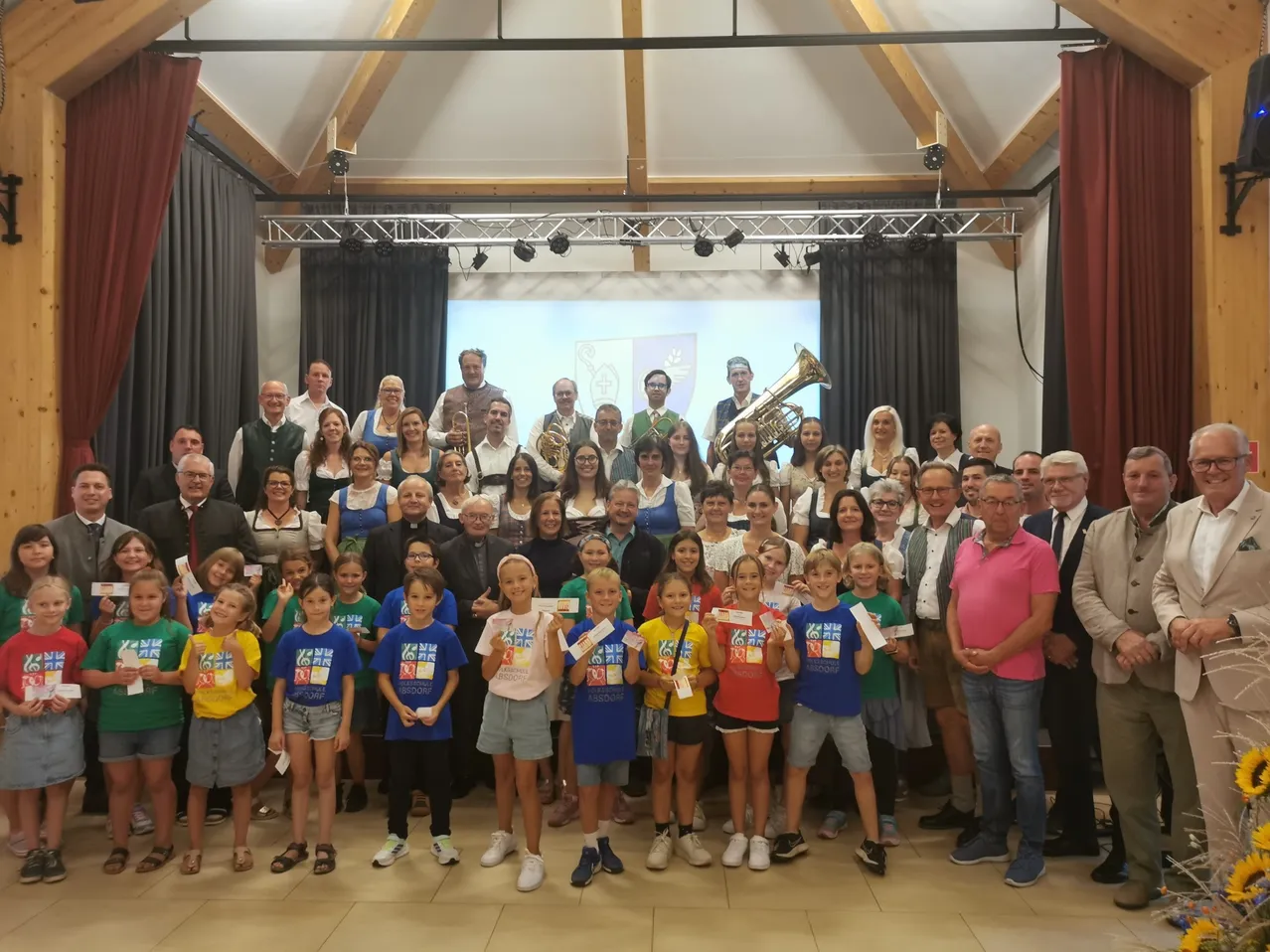 Eine Gruppe von Menschen posiert für ein Foto, einschließlich Erwachsener und Kinder. Einige halten Urkunden. Musikinstrumente stehen hinter ihnen. Eine Projektionsleinwand befindet sich an der Wand.