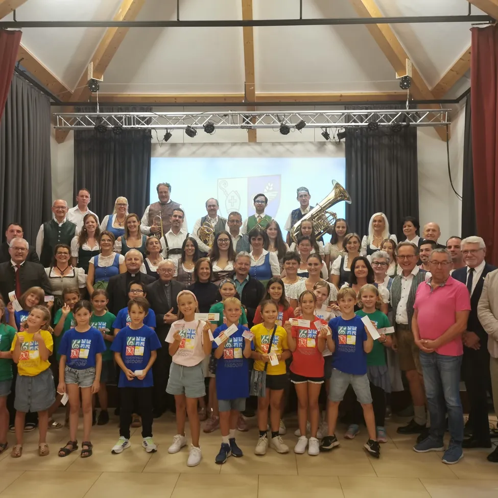 Eine Gruppe von Menschen posiert für ein Foto, einschließlich Erwachsener und Kinder. Einige halten Urkunden. Musikinstrumente stehen hinter ihnen. Eine Projektionsleinwand befindet sich an der Wand.