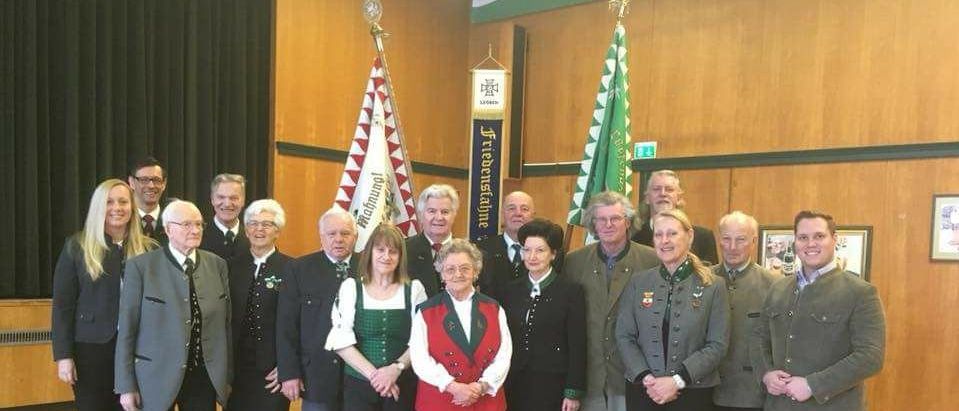 Eine Gruppe von Menschen in traditioneller Kleidung steht vor einer Holzwand mit Fahnen. Dazu gehören Männer und Frauen, einige tragen Brillen und Krawatten. Eine Frau in der Mitte trägt eine rote Weste, und eine Frau neben ihr trägt ein grünes Kleid. Ein Mann auf der linken Seite trägt einen Anzug und Lederschuhe.