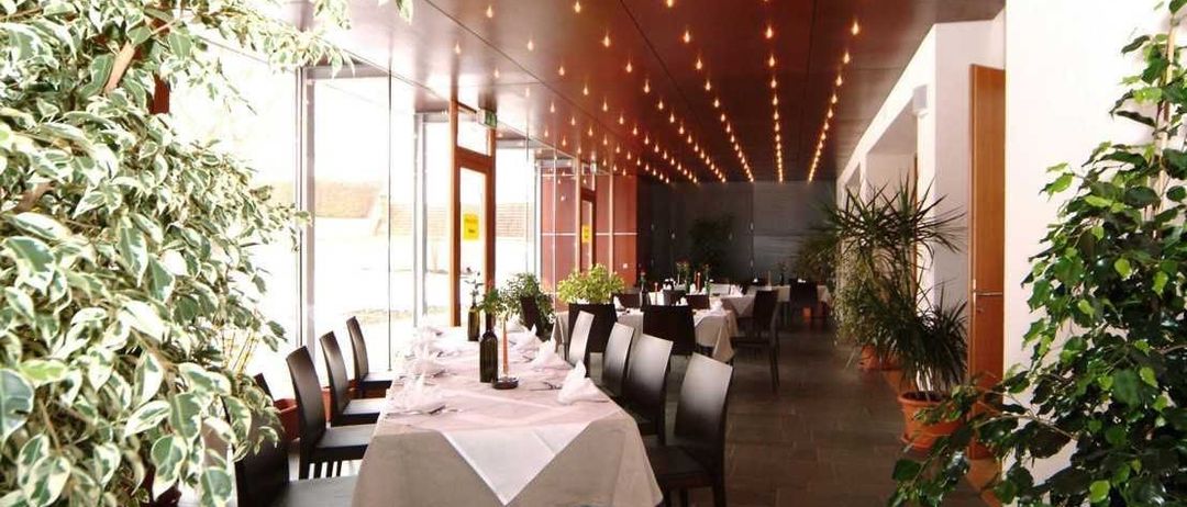 Ein leerer Speisesaal mit langen Tischen, Stühlen und Pflanzen. Die Decke hat eine Reihe kleiner Lichter. Es gibt auch andere Tische mit weißen Tischdecken.