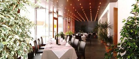 Ein leerer Speisesaal mit langen Tischen, Stühlen und Pflanzen. Die Decke hat eine Reihe kleiner Lichter. Es gibt auch andere Tische mit weißen Tischdecken.