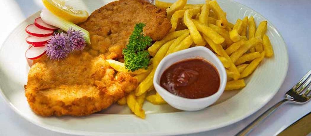 Ein Teller mit einem gebratenen, panierten Fleisch, Pommes Frites, einem kleinen Ketchup-Schälchen, Zitronenscheiben und Radieschen.