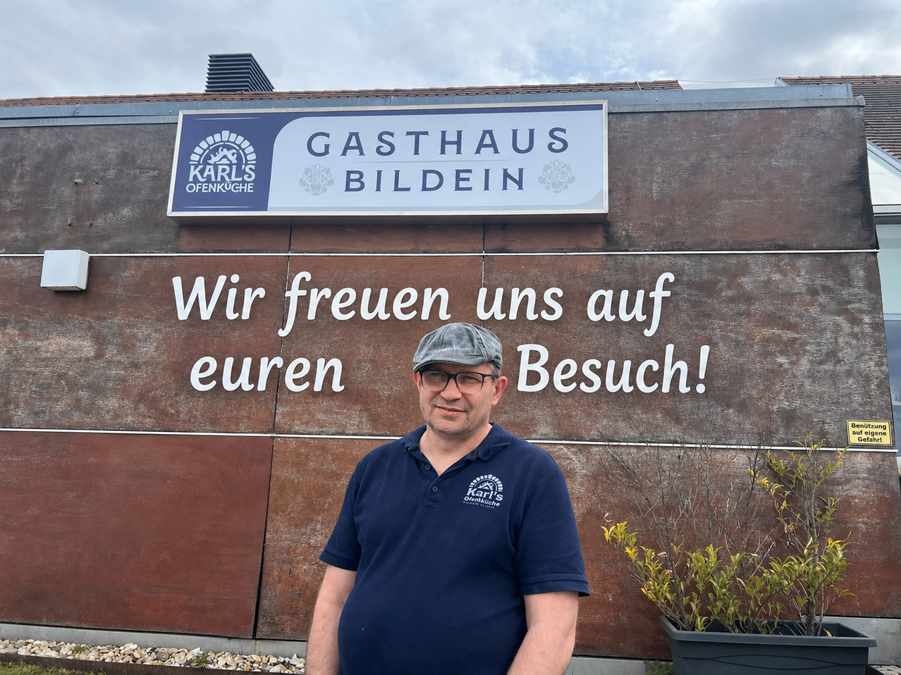 Ein Mann steht vor einem Gebäude mit einem Schild, das 'Gasthaus Bildein' und 'Wir freuen uns auf euren Besuch!' besagt. Er trägt ein blaues Poloshirt und eine Kappe.