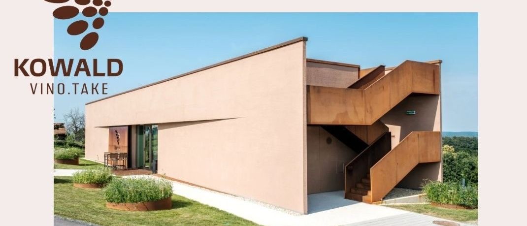 Ein Bild eines modernen Gebäudes mit einem Logo darüber, mit einem beigefarbenen Äußeren und einer braunen Treppe, umgeben von Pflanzen und einem klaren blauen Himmel. Das Gebäude trägt die Aufschrift 'Verkostung in der VINO.TAKE'.