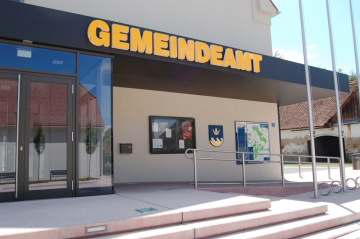 Der Eingang eines Gemeindeamtes mit einer Glastür, gelbem Schild mit der Aufschrift 'GEMEINDEAMT' und einer Karte an der Wand.