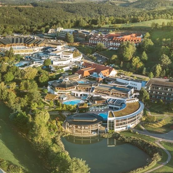 Luftaufnahme eines großen Resortkomplexes mit mehreren Gebäuden, Swimmingpools und üppiger Grünfläche. Im Vordergrund ein kreisförmiges Gebäude mit Pool.