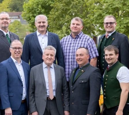 Eine Gruppe von Männern und Frauen in formeller Kleidung, einschließlich Anzüge und Krawatten, posiert für ein Foto in einem Park mit Bäumen und einem Gebäude im Hintergrund.