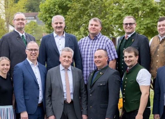 Eine Gruppe von Männern und Frauen in formeller Kleidung, einschließlich Anzüge und Krawatten, posiert für ein Foto in einem Park mit Bäumen und einem Gebäude im Hintergrund.