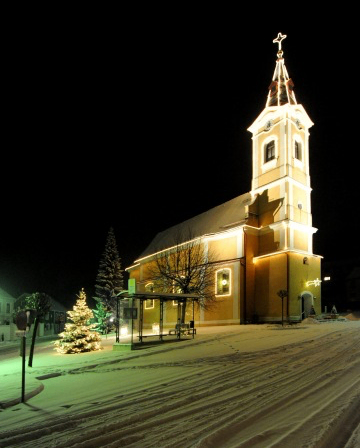 Eine Kirche mit einem Turm und einem Kreuz ist nachts beleuchtet, umgeben von Schnee und einem Weihnachtsbaum.