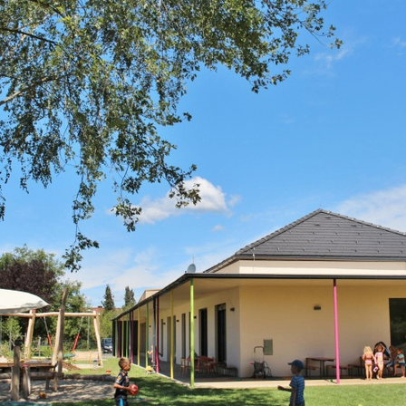 Ein Gebäude mit grauem Dach und grüner Einfassung. Kinder spielen auf einer Rasenfläche mit einem Baum und einem Baldachin.