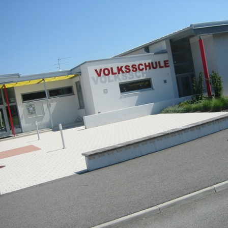Ein weißes Gebäude mit dem Wort 'Volksschule' in Rot auf der Seite. Der Eingang hat ein gelbes Vordach.