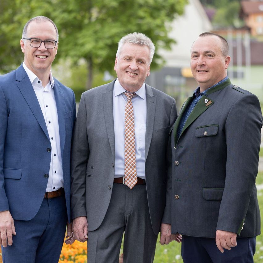 Drei Männer in Anzügen und Krawatten stehen zusammen in einem Park. Der Mann auf der linken Seite trägt einen marineblauen Anzug, der Mann in der Mitte einen grauen Anzug und der Mann auf der rechten Seite trägt eine grüne Uniform mit Knöpfen.