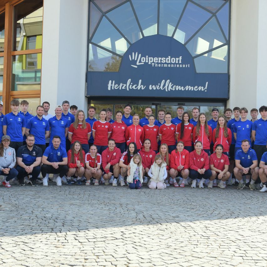 Eine Gruppe von Athleten posiert für ein Foto vor einem Gebäude mit einem Schild, das 'Loipersdorf Thermeneresort Herzlich willkommen!' steht. Sie sind alle in passenden roten und blauen Uniformen gekleidet.