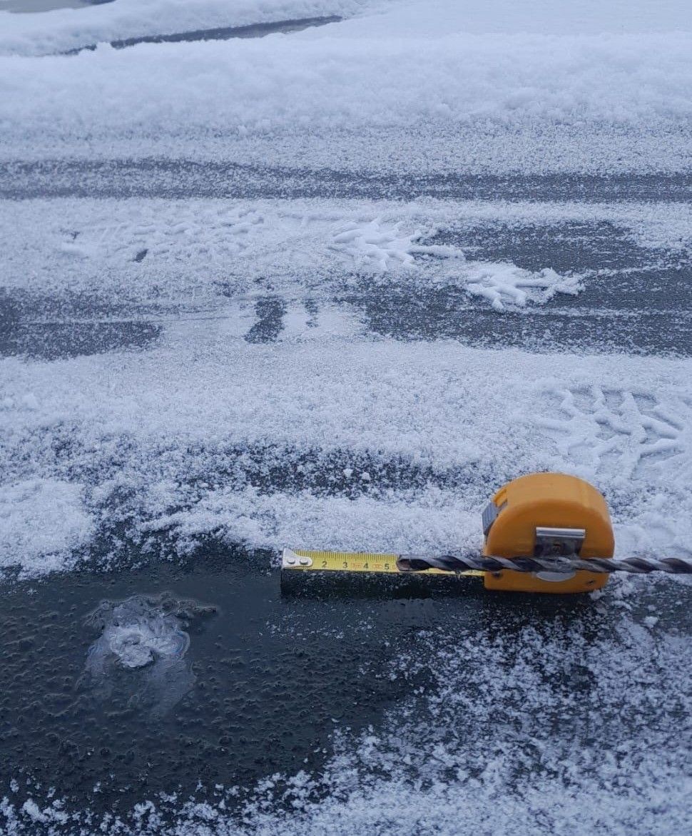 Ein gelbes Messband liegt auf einer verschneiten Oberfläche mit sichtbaren Reifenspuren und einer Pfütze. Der Schnee ist teilweise geschmolzen und zeigt den Boden.