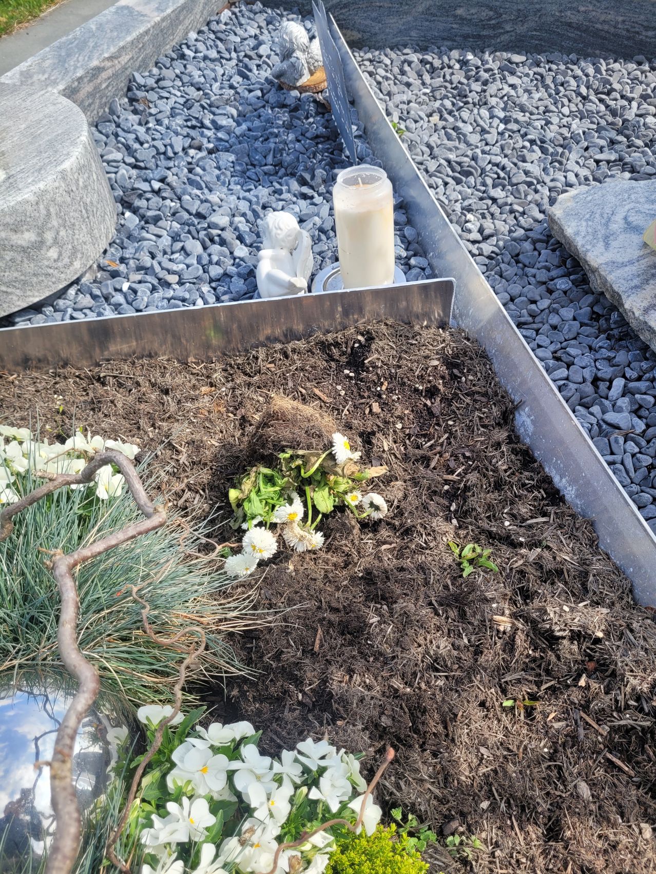 Ein Gartenbeet mit brauner Erde und kleinen weißen Blumen ist zu sehen. Ein kleines Kerze und eine weiße Engelstatue sind in der Nähe platziert.