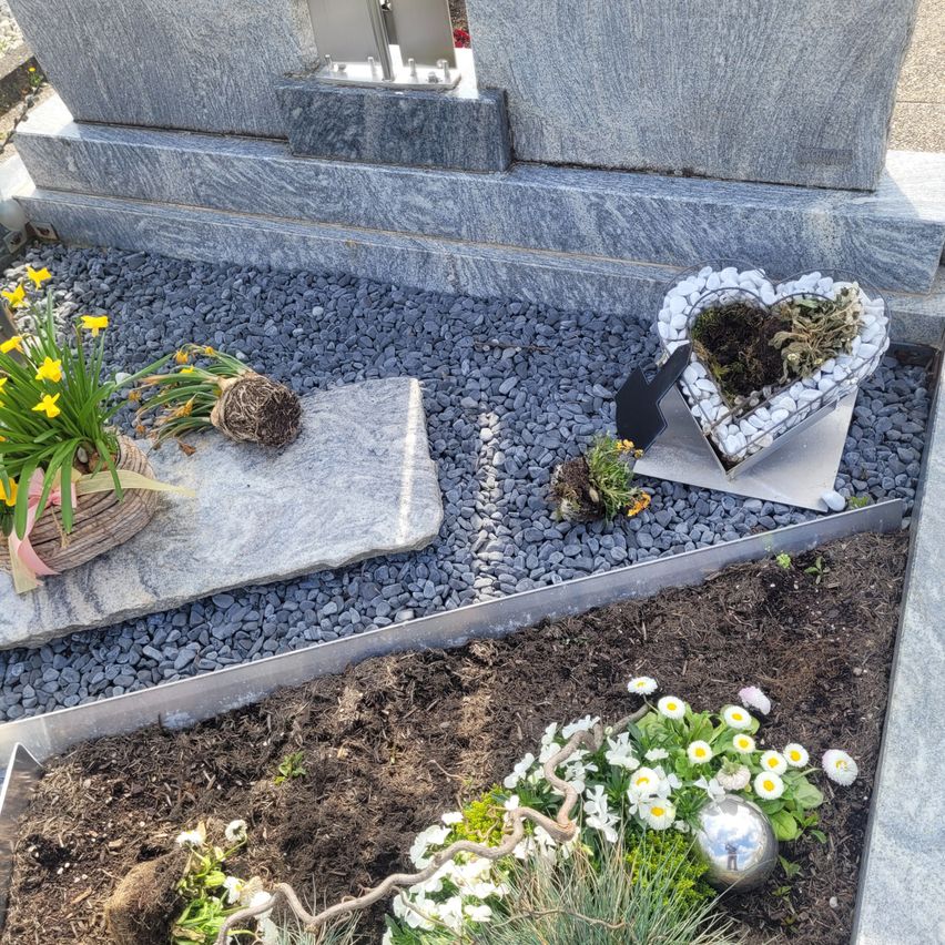 Ein Grab mit einem herzförmigen Pflanzgefäß, einer silbernen Kugel und weißen Blumen auf einem Kiesweg.