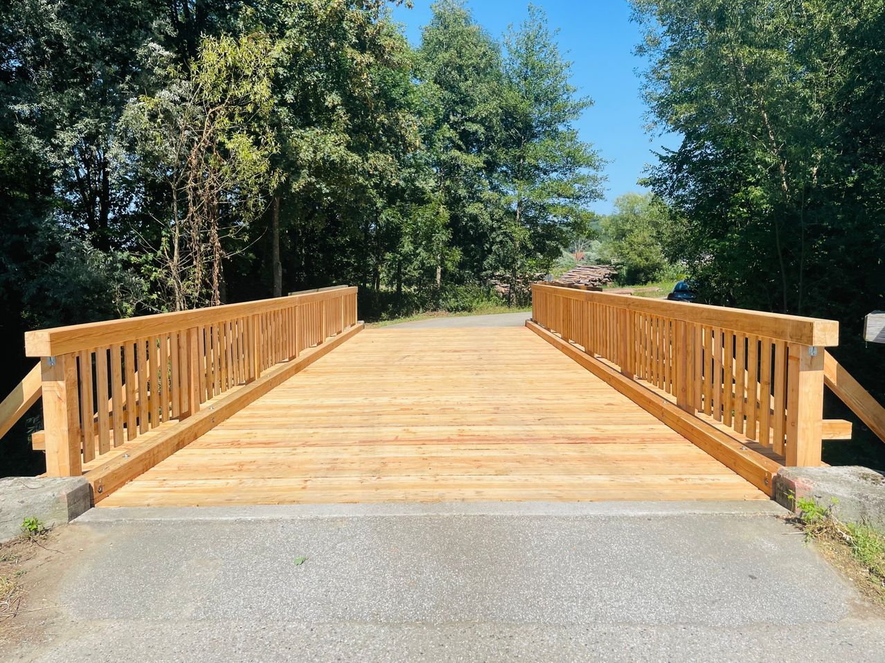 Eine Holzbrücke mit Geländer überspannt eine Straße, umgeben von üppigem Grün unter einem klaren blauen Himmel.