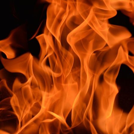Eine Nahaufnahme eines Feuers mit orangen und gelben Flammen. Die Flammen sind wellig und dynamisch, gegen einen dunklen Hintergrund beleuchtet.